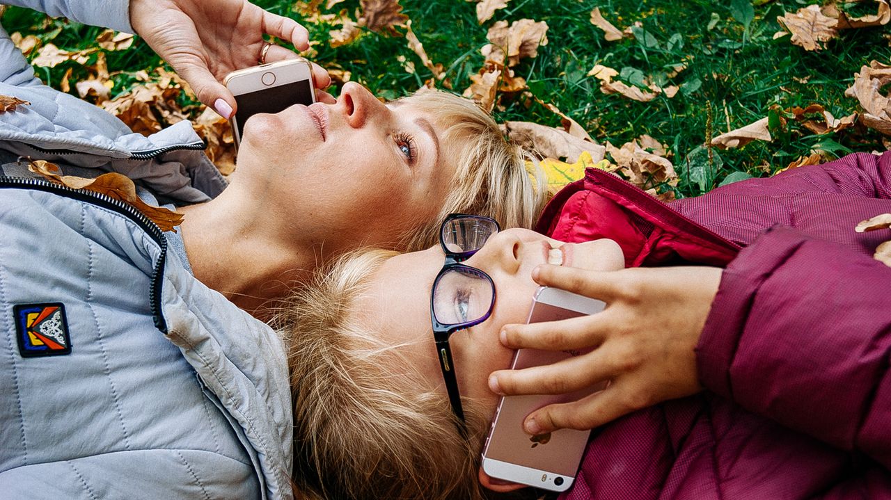A blonde mother and her son lying on the grass, using mobile phones together outdoors.