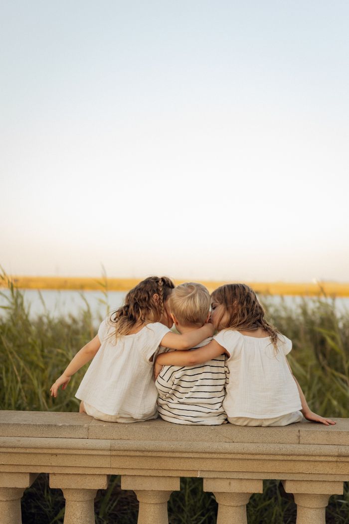 about-02 Three children sitting on a stone railing, embracing by the water at sunset.
