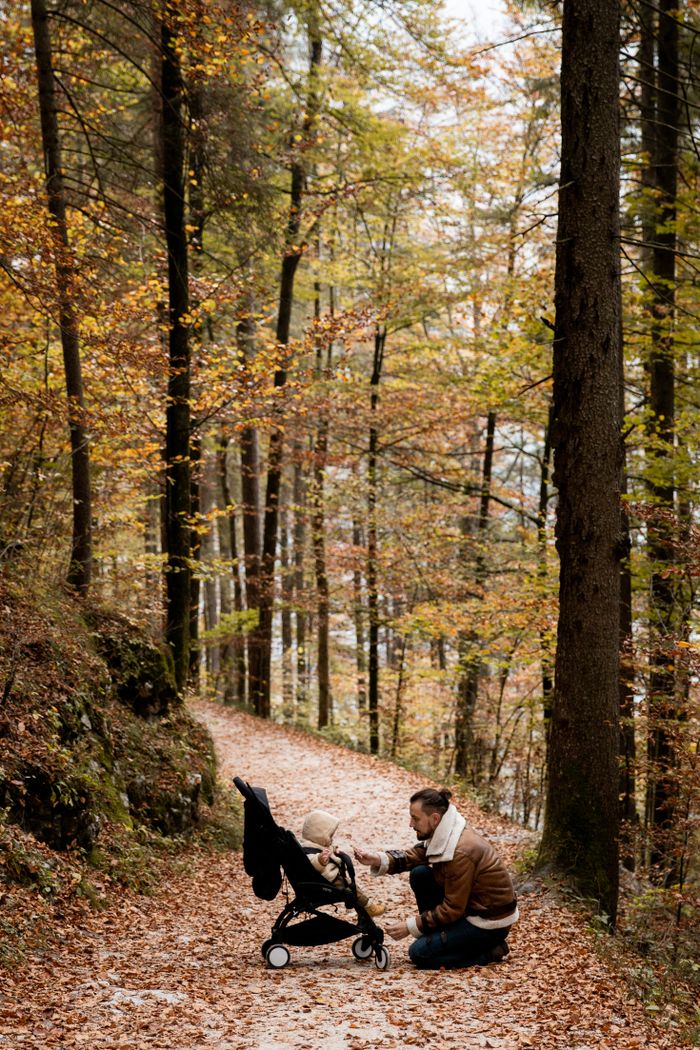 ours-journey A father kneels with his toddler in a stroller along a wooded path in fall.
