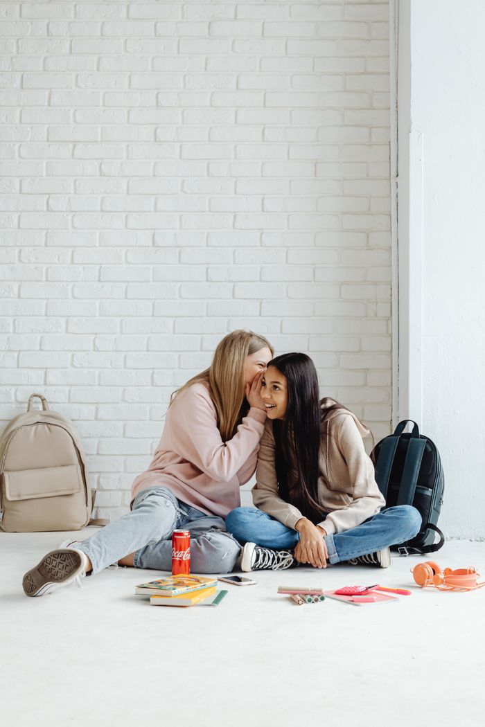 Two teenage girls whispering, surrounded by school supplies in a bright, indoor setting.